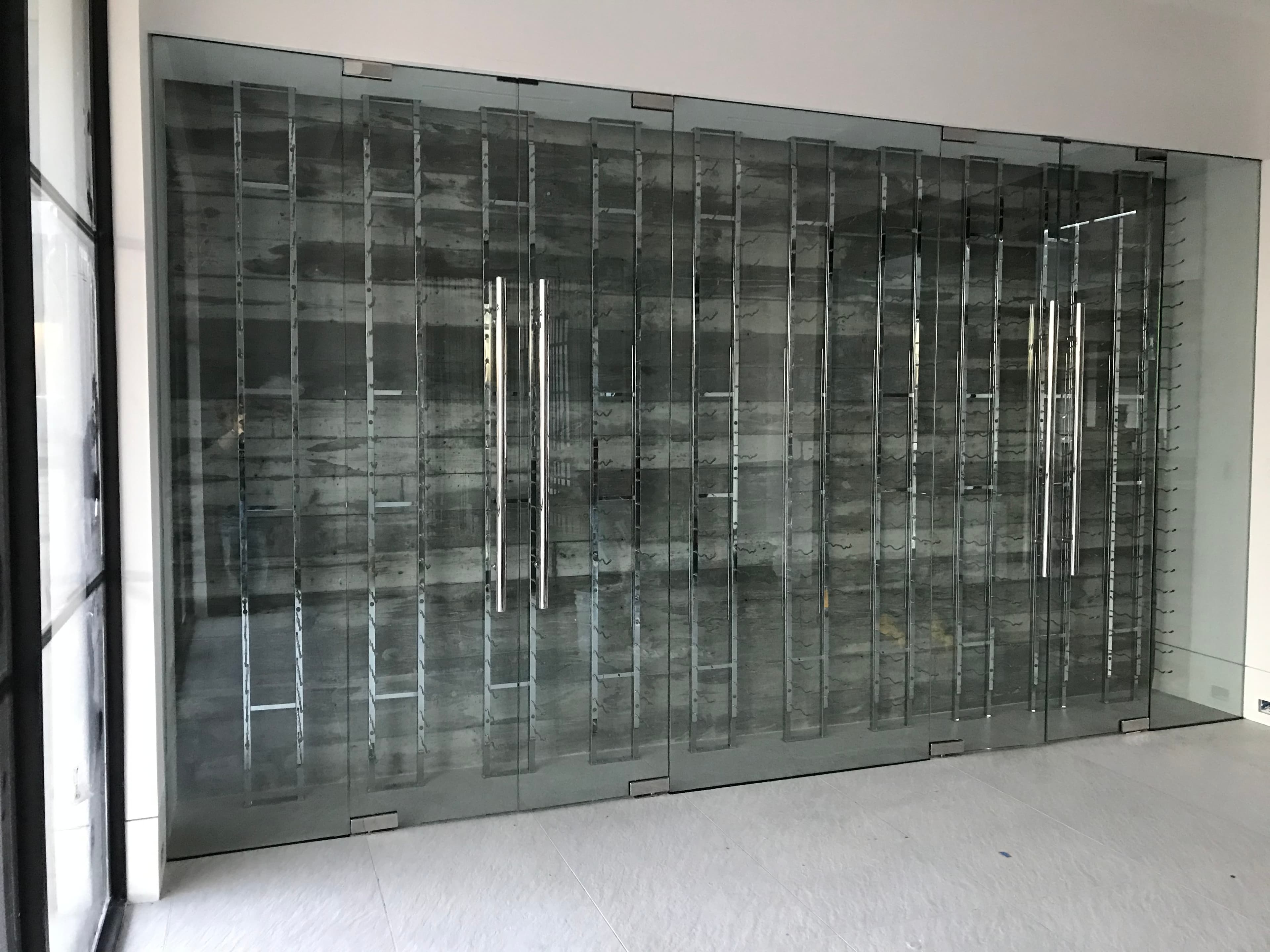 Modern glass-enclosed wine cellar with vertical metal racks and a textured grey back wall.