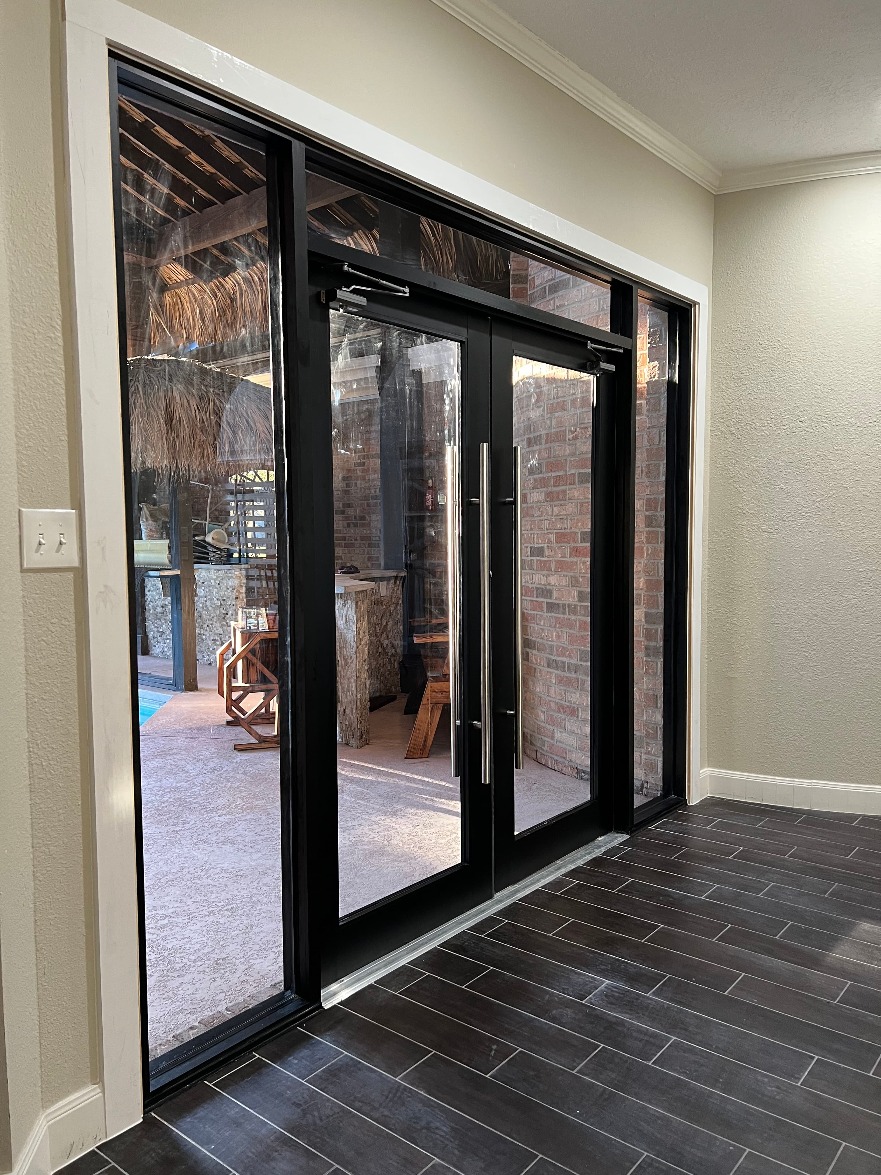 Black-framed glass double doors with silver handles lead to an outdoor patio with a thatched roof.