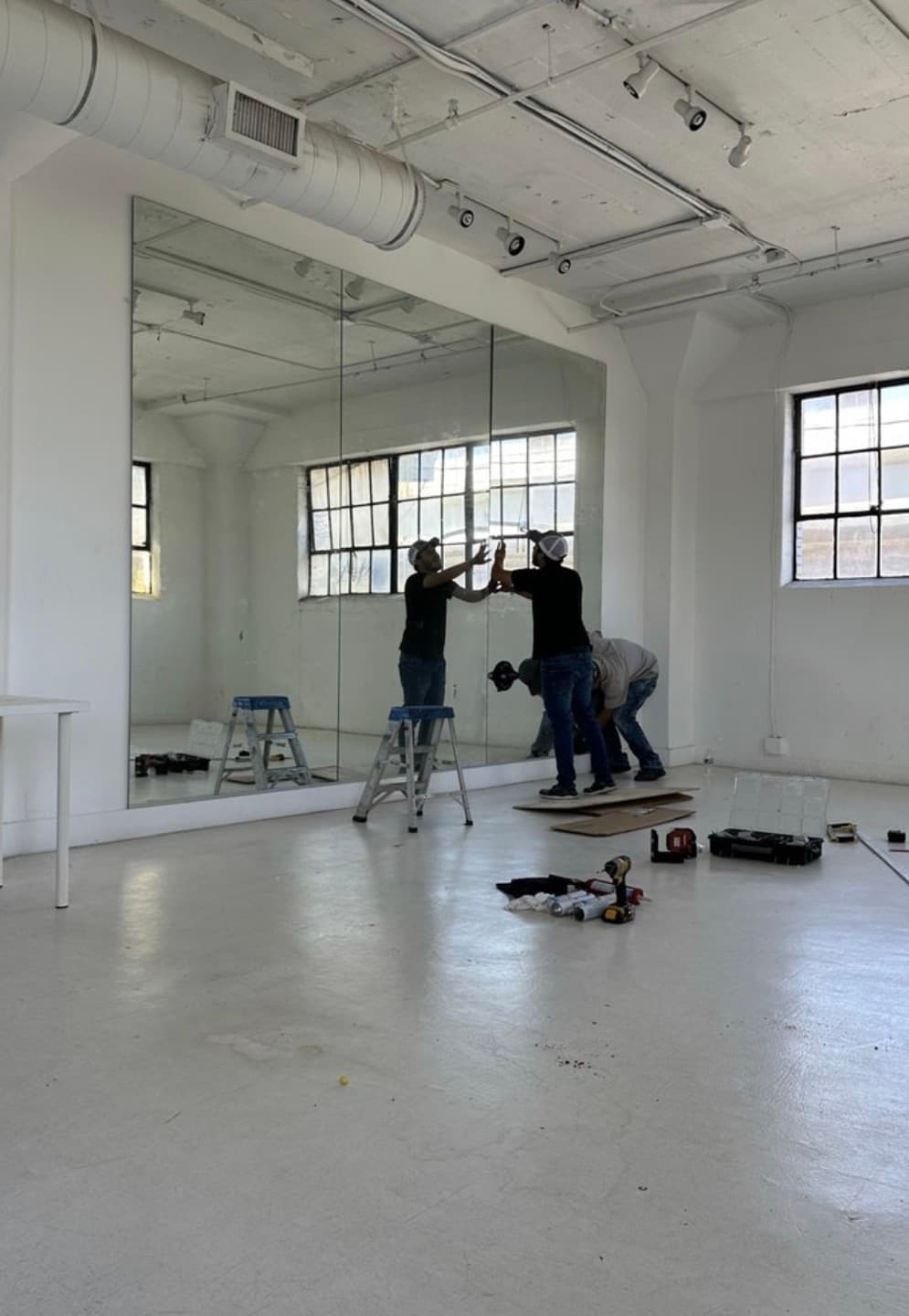 Two workers mounting large mirrors in a bright white studio with tools and ladders.