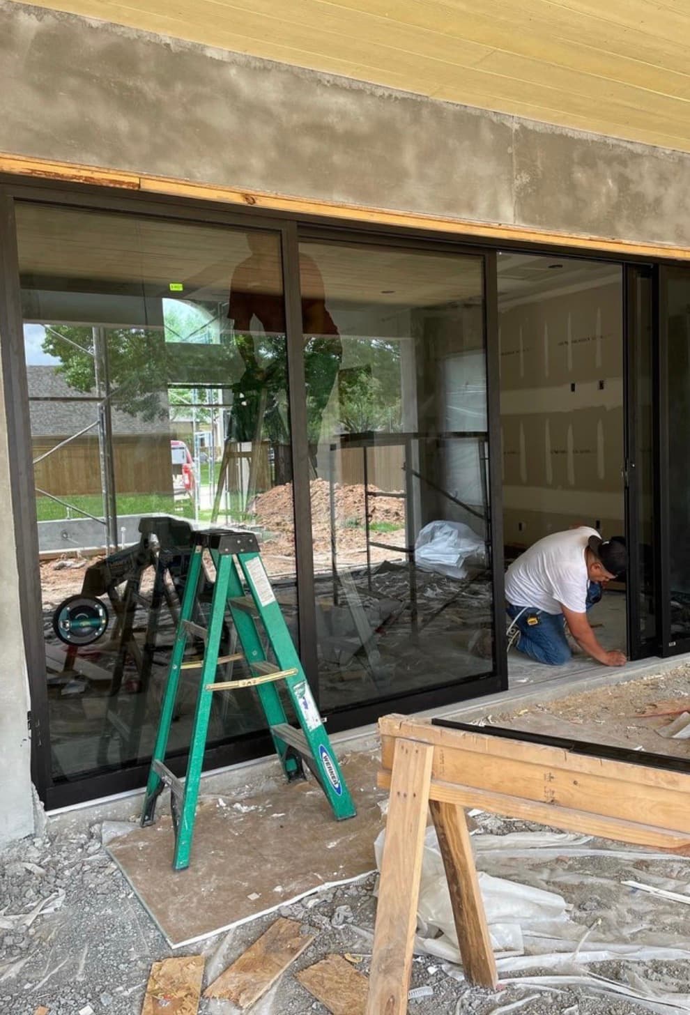 Worker installs a large black-framed sliding glass door at a construction site with a ladder.