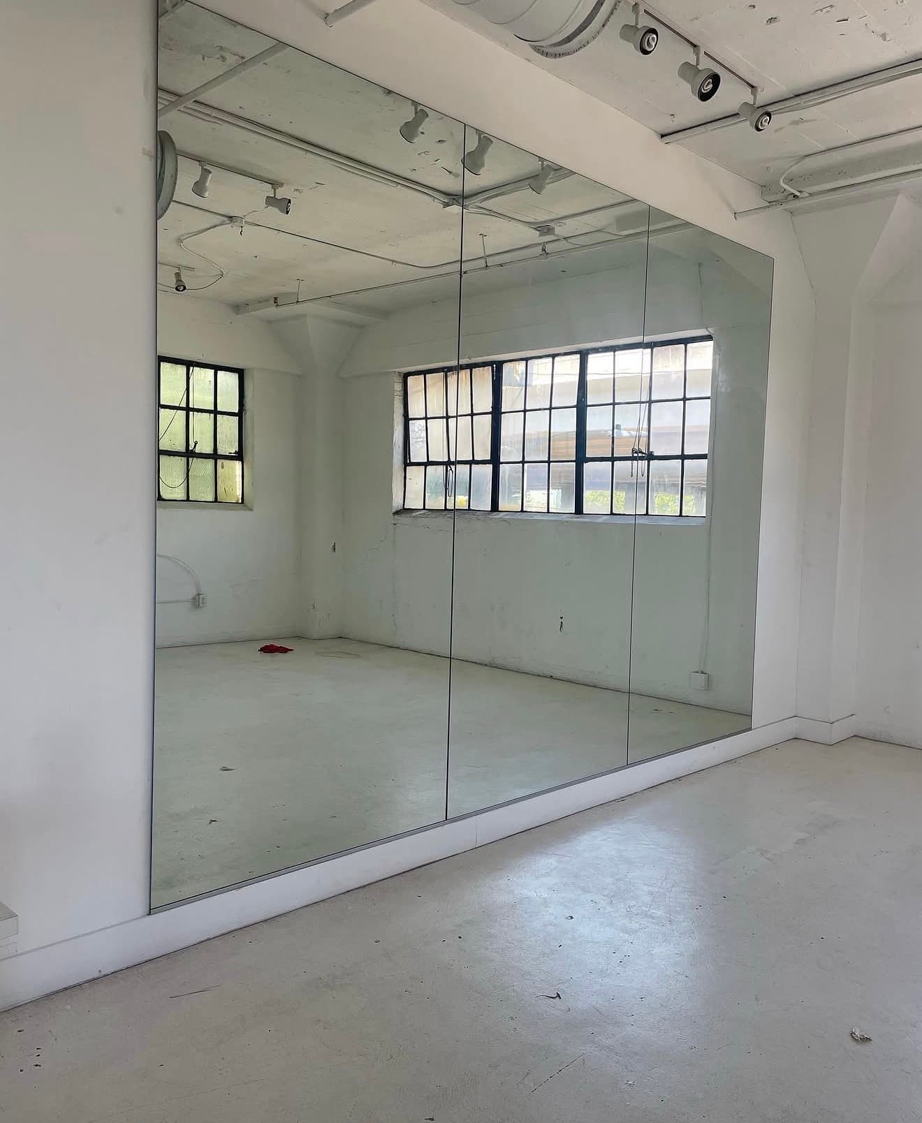 Large wall mirror in an empty white studio reflecting industrial windows and track lighting.