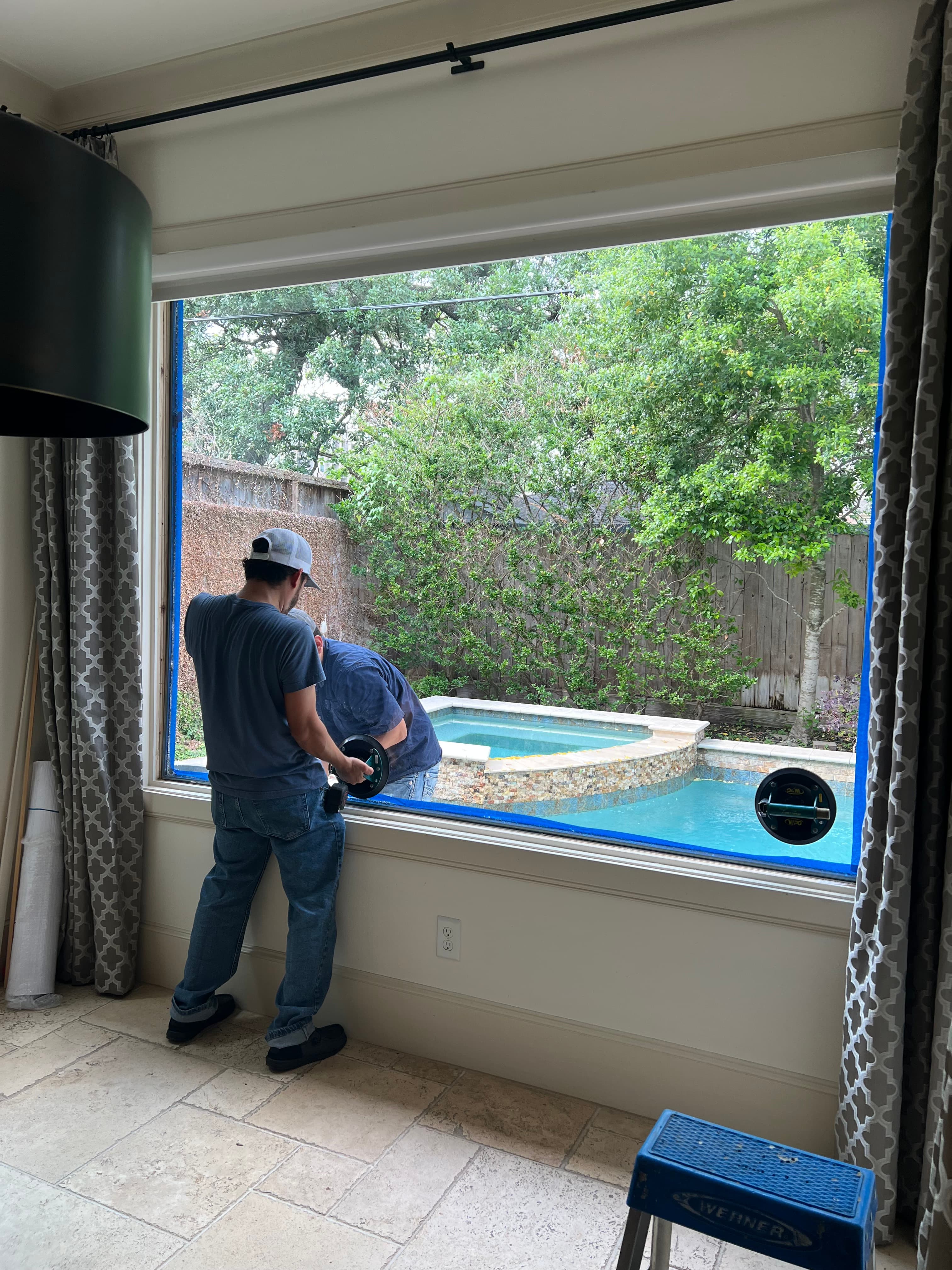 Two workers use suction cups to install a large window overlooking a backyard pool.