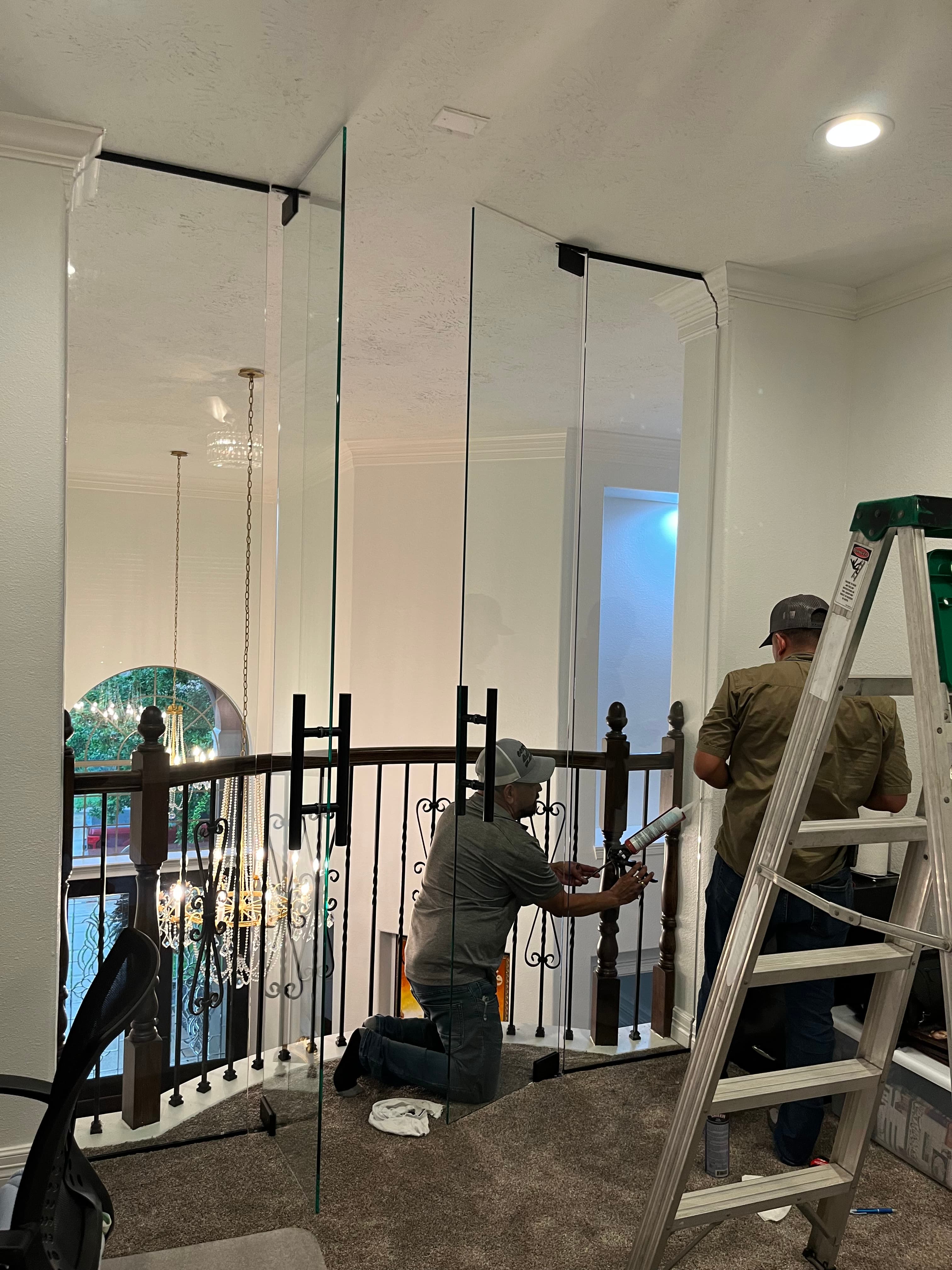 Two workers installing tall glass partitions with black handles on a second-floor home landing.