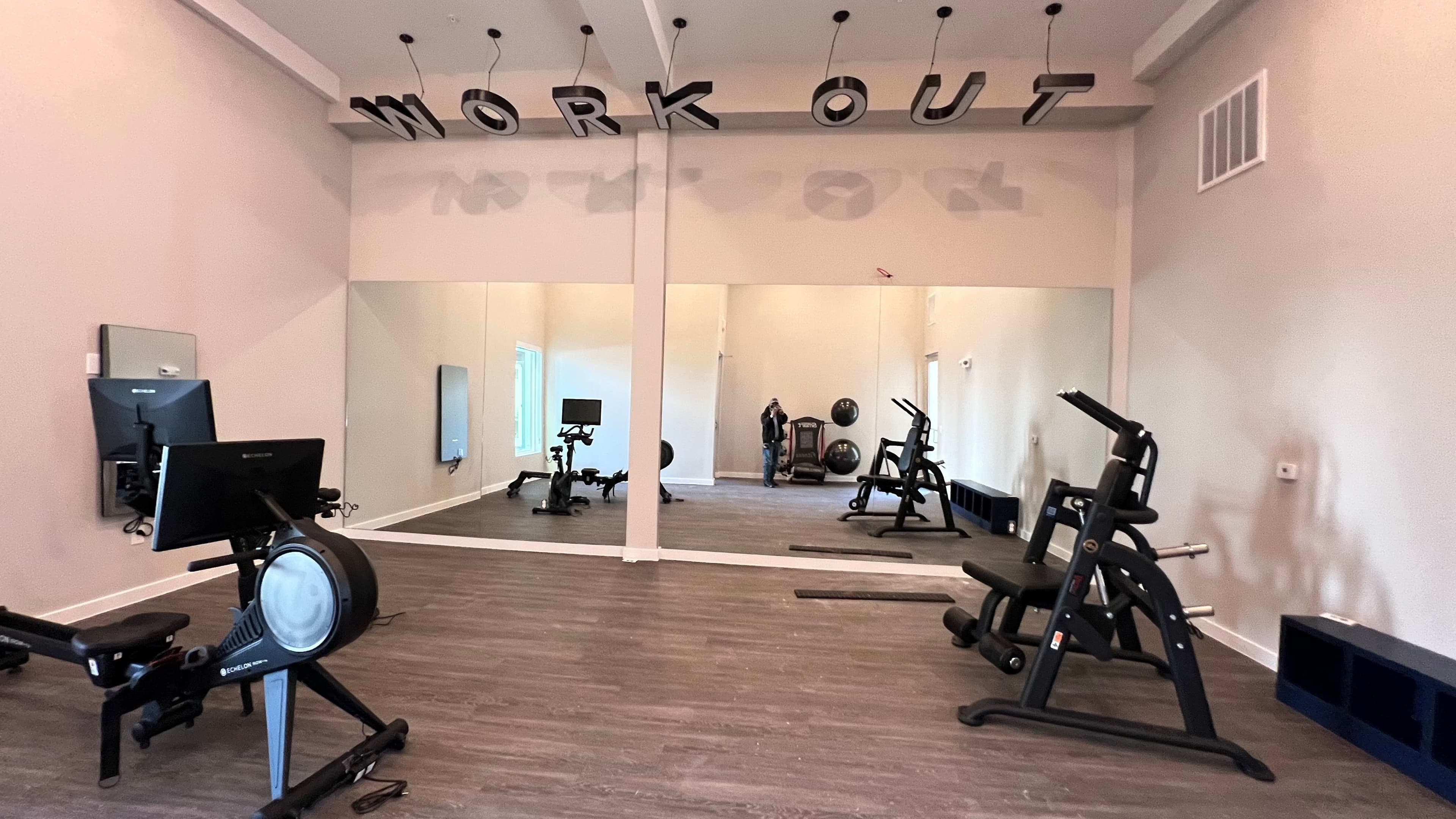 Modern gym with exercise equipment, large wall mirrors, and a hanging sign reading 'WORK OUT'.